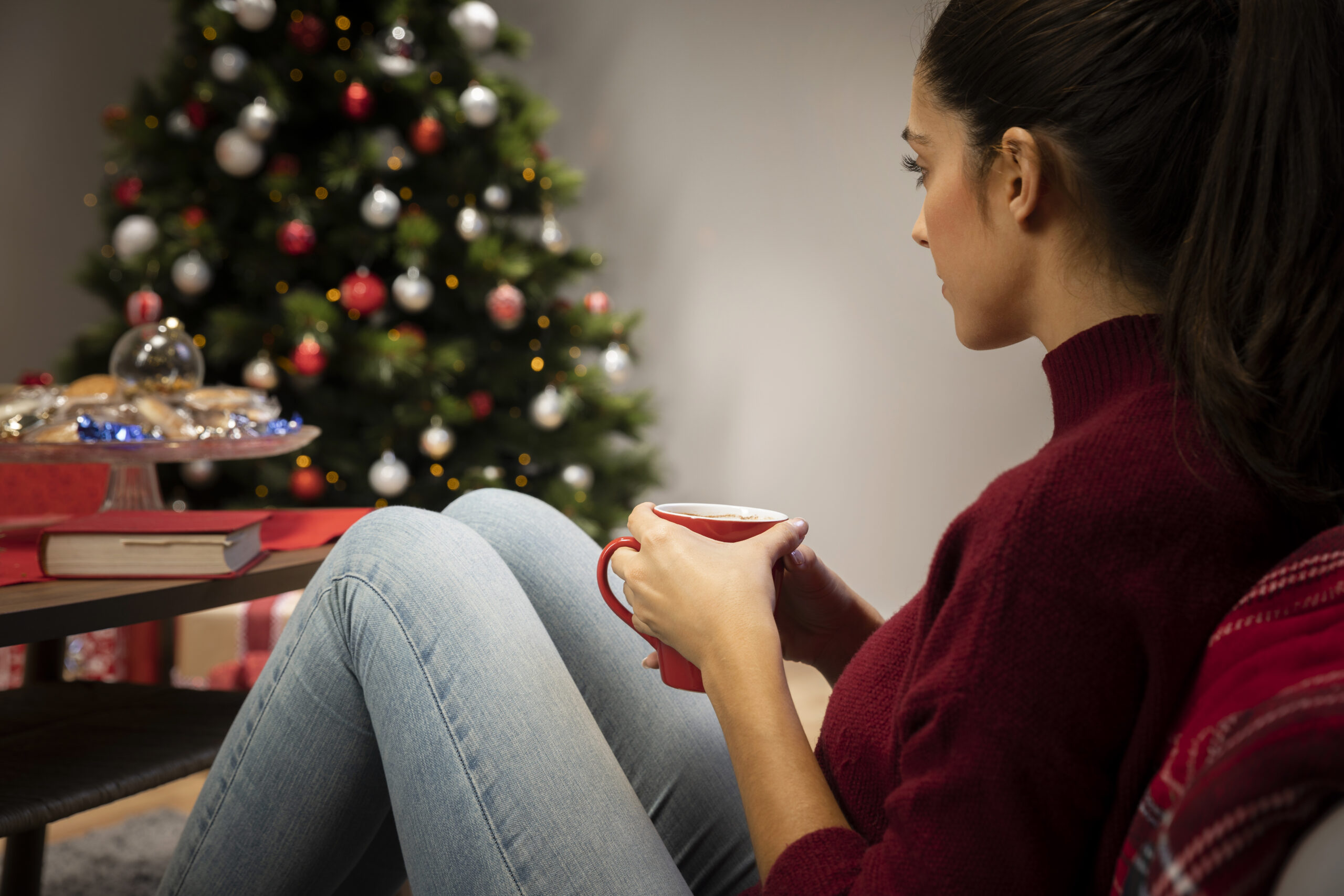Woman sitting quietly with a mug near a Christmas tree, reflecting during the holiday season and symbolizing depression during the Christmas season