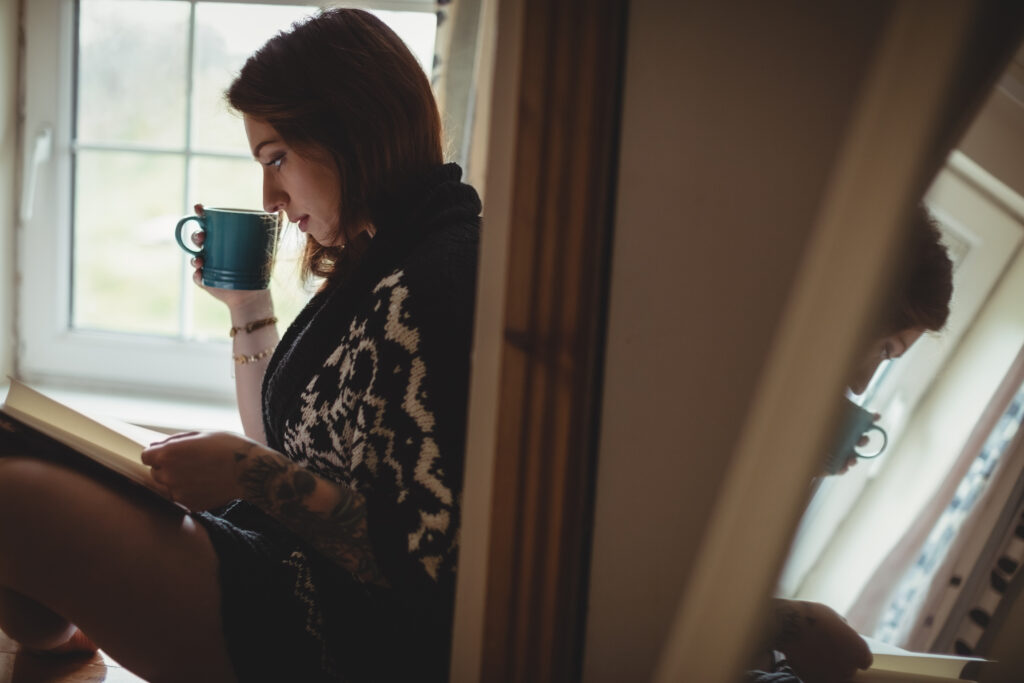 Woman sitting by a window reading a book and holding a warm mug, creating a quiet moment to calm her nervous system and ease everyday anxiety.