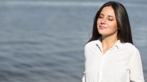 Woman standing by the water with eyes closed, breathing deeply as sunlight and breeze help calm her nervous system and reduce everyday anxiety.