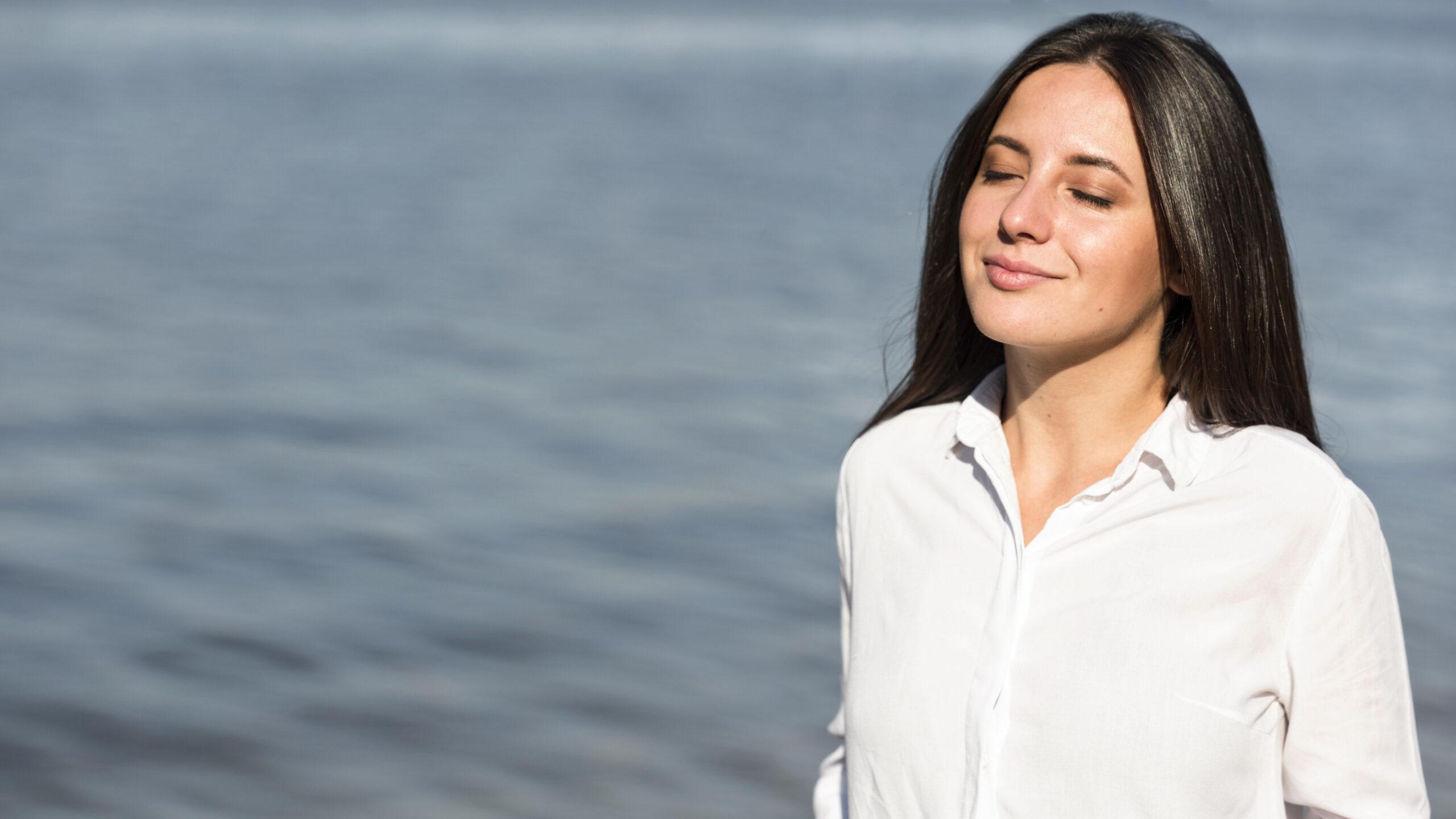 Woman standing by the water with eyes closed, breathing deeply as sunlight and breeze help calm her nervous system and reduce everyday anxiety.
