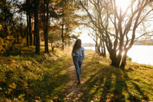 Woman walking slowly along a wooded path in autumn sunlight, practicing grounding to calm her nervous system and ease everyday anxiety triggers.
