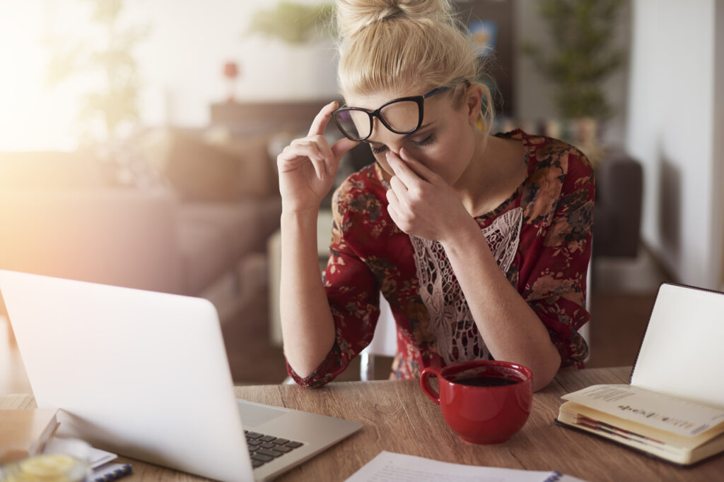 Woman experiencing emotional burnout sitting at a desk with a laptop, holding her glasses and rubbing her eyes from exhaustion